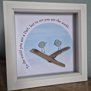 May include: White framed artwork with a blue and white background. Two small, gray pebble birds are perched on a piece of driftwood. The text reads, "To the world you are a Dad, but to me you are the world."