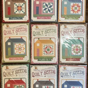 May include: Nine quilt pattern books titled "Quilt Seeds" are displayed. Each book cover features a house illustration with a unique quilt block design. The color palette includes red, blue, green, and brown. The text "Neighbor No. 1" through "Neighbor No. 9" is visible.