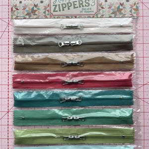May include: A package of eight 16-inch zippers in various colors, including white, gray, brown, red, teal, green, and light blue. The package is labeled "Happy Zippers 3" and includes text from "Lori Holt of Bee in my Bonnet."