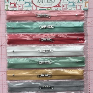May include: A package of Happy Zippers in various colors, including pink, teal, red, white, green, gold, gray, and turquoise. Each zipper is individually packaged and features a silver metal pull. The packaging has a sewing machine pattern and the text "Happy Zippers".