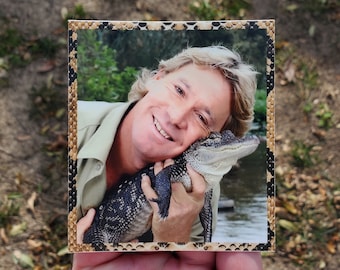 May include: A square print featuring a smiling man holding a small alligator. The print has a decorative border with a snakeskin pattern. The man is wearing a khaki shirt.