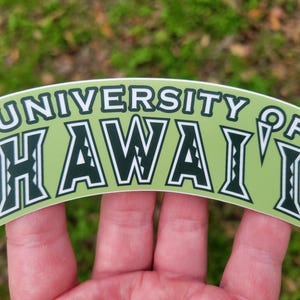 May include: A curved sticker featuring the text UNIVERSITY OF HAWAI'I in dark green letters with white outlines on a light green background. The letters have a decorative geometric pattern inside them, and the sticker has a white border.
