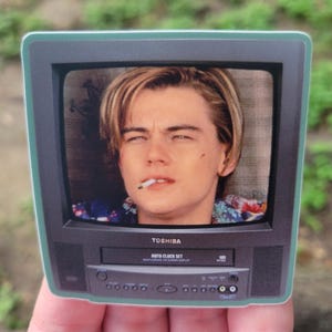 Peut inclure: Un autocollant rectangulaire en forme de t&eacute;l&eacute;viseur noir vintage Toshiba avec magn&eacute;toscope int&eacute;gr&eacute;. L'&eacute;cran affiche le portrait d'une jeune personne aux cheveux blonds avec une cigarette &agrave; la bouche. Le t&eacute;l&eacute;viseur porte le logo Toshiba.
