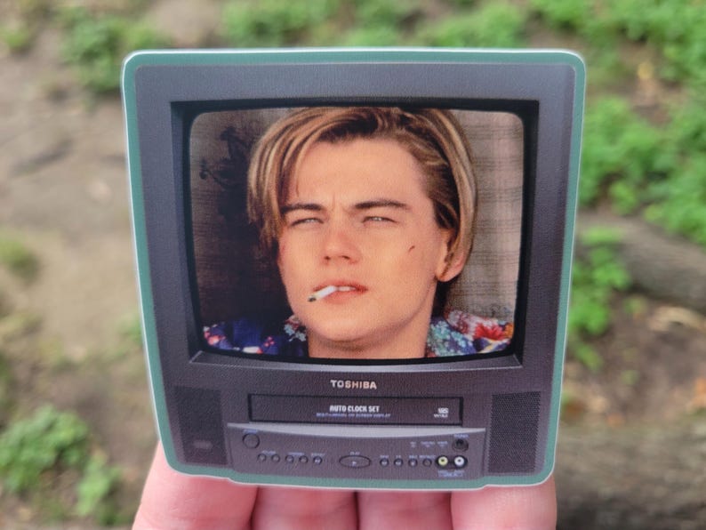 Peut inclure: Un autocollant rectangulaire en forme de t&eacute;l&eacute;viseur noir vintage Toshiba avec magn&eacute;toscope int&eacute;gr&eacute;. L'&eacute;cran affiche le portrait d'une jeune personne aux cheveux blonds avec une cigarette &agrave; la bouche. Le t&eacute;l&eacute;viseur porte le logo Toshiba.