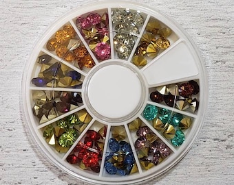 May include: A white, circular container with multiple compartments filled with colorful nail art rhinestones. The rhinestones are various shapes and colors, including gold, red, blue, green, and pink. The container is on a white, textured surface.