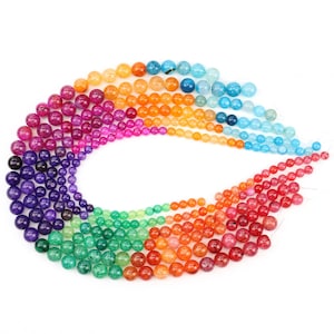 May include: A collection of colorful gemstone beads arranged in a rainbow arc. The beads range in size and color, from deep purple to light blue, including shades of green, orange, and red. The beads are round and appear to be strung together.
