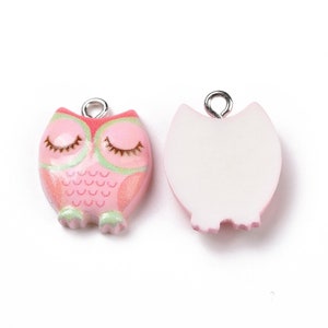 May include: Two owl-shaped charms. One charm is pink with green and red accents, closed eyes, and a silver loop. The other charm is white with a pink outline and a silver loop. The charms are likely for jewelry making.