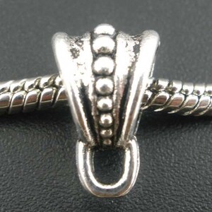 May include: A silver-toned charm with a decorative design, featuring a series of small, raised spheres and a loop at the bottom. The charm is attached to a silver chain, and the background is dark.