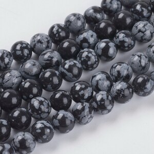 May include: A collection of round, black snowflake obsidian beads. Each bead has a polished, glossy finish and features a unique pattern of white or gray snowflake-like inclusions. These beads are strung together, ready for jewelry making.