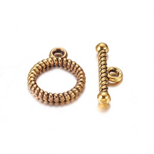 May include: A pair of gold-colored toggle clasps. One clasp is a ring with a textured, twisted design. The other is a bar with a similar twisted texture and a small loop. Both clasps have a vintage aesthetic.