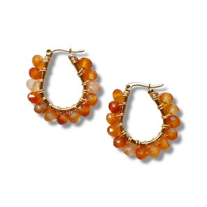 May include: A pair of gold-colored hoop earrings. Each earring features a teardrop shape with a cluster of orange and peach-colored gemstone beads. The beads are wire-wrapped around the hoop.