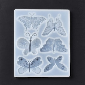 May include: A clear, rectangular silicone mold with six butterfly designs. The butterflies vary in shape and detail, with some featuring intricate patterns. The mold is designed for crafting and creating decorative elements.