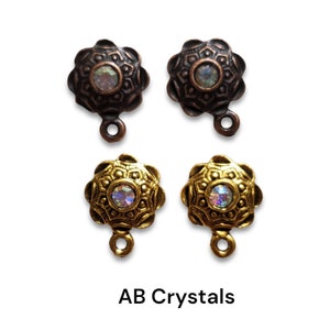 May include: Four ornate metal jewelry components. Two are bronze-colored, and two are gold-colored. Each has a central iridescent crystal and a loop for attachment. The text "AB Crystals" is at the bottom.