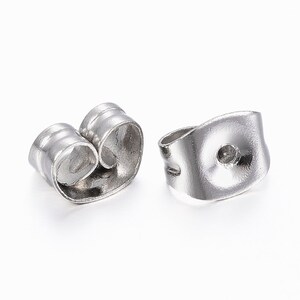 May include: Two silver-colored earring backs on a white background. The earring backs are made of a shiny metal and have a curved design. One back is in focus, showing a small hole for the earring post.