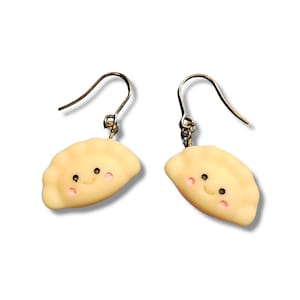 May include: A pair of dangle earrings featuring smiling dumpling charms. The charms are light yellow with pink cheeks and black eyes. The earrings have silver-colored fish hook ear wires.