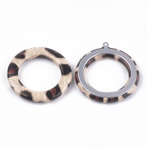 May include: Two round jewelry components with a leopard print design. One component is a complete ring, while the other has a silver-colored metal frame with a loop at the top. The leopard print features black, brown, and tan colors.
