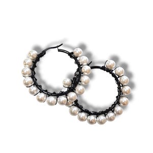 May include: A pair of hoop earrings featuring faux pearls and black wire. The earrings have a circular design with the pearls evenly spaced around the hoops. The pearls are white and the wire is black.