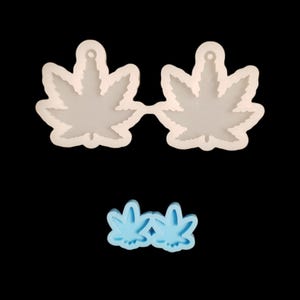 May include: Two pairs of cannabis leaf-shaped molds, one in white and the other in light blue, are displayed against a black background. The molds are likely for crafting earrings or pendants, showcasing a botanical theme.