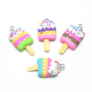 May include: Four colorful popsicle charms with a white frosting-like topping and rainbow sprinkles. Each charm has a light yellow stick and a silver loop for attaching to jewelry or other crafts. The popsicles are pink, green, yellow, and brown.