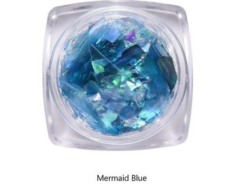 May include: A clear, square container filled with iridescent blue and green flakes. The flakes are labeled "Mermaid Blue" and appear to be for nail art or craft projects. The flakes have a shimmering, reflective quality.