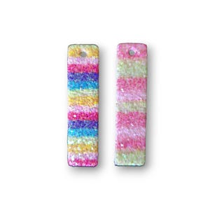 May include: Two rectangular, glittery pendants. One features horizontal stripes of yellow, blue, pink, and purple. The other has pink and yellow stripes. Each has a small hole at the top for stringing. These are likely jewelry components.