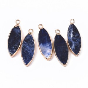 May include: Five elongated, oval-shaped pendants with a dark blue and white marbled stone center, framed by a gold-colored metal. Each pendant has a small loop at the top for stringing. The pendants are displayed against a white background.
