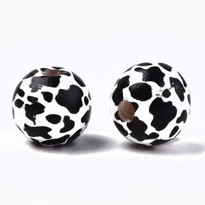 May include: Two round wooden beads with a cow print design. The beads are white with irregular black spots. Each bead has a hole drilled through the center. These beads are suitable for jewelry making and crafts.