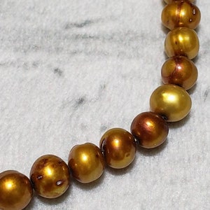 May include: A close-up of a strand of golden-colored pearls. The pearls have a lustrous, iridescent sheen, with variations in color from light gold to deep bronze. The pearls are round and evenly sized, suggesting a necklace or bracelet.