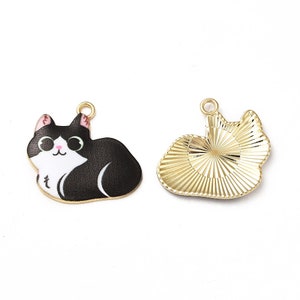 May include: Two cat-shaped charms. One is a black and white enamel cat with a gold-colored loop. The other is a gold-colored charm with a textured, radiating design, also in the shape of a cat. Both charms are on a white background.