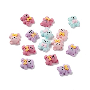 May include: A collection of small, colorful bear-shaped embellishments. The bears are in shades of pink, purple, and blue, each with a yellow bow. These decorative pieces are scattered on a white surface.