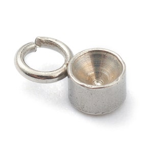 May include: A close-up of a silver-colored jewelry finding, featuring a small, open ring connected to a cylindrical cup-shaped component. The cup has a conical interior and a small hole at the bottom.