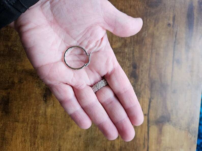 Peut inclure: Une boucle d'oreille argent&eacute;e avec une fermeture &agrave; charni&egrave;re. La boucle d'oreille est tenue dans une main ouverte, avec une bague au doigt. Le fond est une surface en bois.