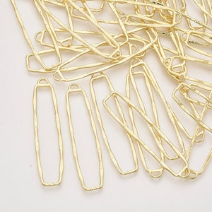 May include: A collection of gold-colored, rectangular metal jewelry components. Each component has a textured surface and a small loop at one end. The components are scattered across a light background.