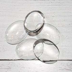 Puede incluir: Un conjunto de cabujones de vidrio transparente de forma ovalada. Un cabujón está apilado encima de los demás. Los cabujones transparentes reflejan la luz, creando una apariencia brillante y limpia. El fondo es una superficie texturizada de color gris claro.