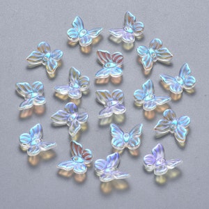 May include: Iridescent butterfly-shaped embellishments scattered across a gray surface. The butterflies have a translucent quality with hints of blue, purple, and gold. These decorative elements are ideal for crafts and jewelry making.