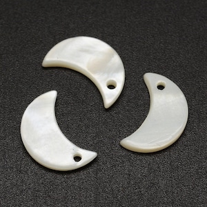 May include: Three white, crescent-shaped mother-of-pearl pendants with a pearlescent sheen. Each pendant has a small hole for stringing. The pendants are arranged on a dark gray surface.