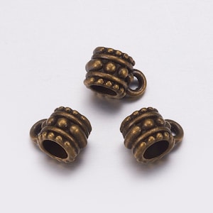 May include: Three antique bronze-colored metal beads. Each bead has a cylindrical shape with a small handle and is decorated with raised circular and dotted patterns. The beads are likely for jewelry making or crafting.