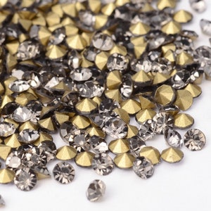 May include: A pile of faceted rhinestones in shades of gray and gold. The rhinestones have a metallic finish and are scattered across a white surface. The gemstones are small and have a flat back, suitable for embellishing various crafts.