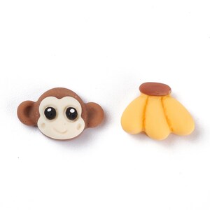 May include: Two small, colorful resin charms. One is a brown monkey face with a cream-colored face and large black eyes. The other is a bunch of yellow bananas with a brown stem. These charms are ideal for crafting.
