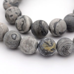 May include: Close-up of a strand of gray and black marbled stone beads. The beads are round and have a matte finish. The beads are strung together, and the background is white.