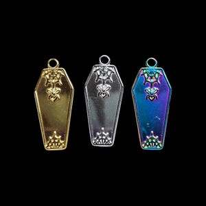 May include: Three coffin-shaped pendants in gold, silver, and iridescent finishes. Each pendant features a decorative spiderweb and floral design. The pendants have a loop at the top for attaching to a chain or other jewelry.