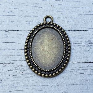 May include: An antique brass-toned oval pendant setting with a beaded border. The setting has a loop at the top for attaching to a chain or cord. The center is recessed, ready for a cabochon or other embellishment. Ideal for jewelry making.