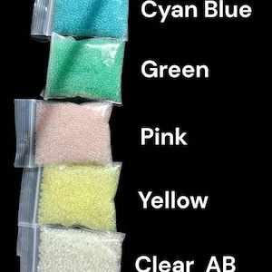 May include: Five clear plastic bags filled with small, colorful beads. The colors include cyan blue, green, pink, yellow, and clear AB. Text on the image reads "Daylight", "Cyan Blue", "Green", "Pink", "Yellow", and "Clear AB".