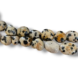 May include: A strand of dalmatian jasper beads. The beads are oval-shaped, with a cream-colored base and black and orange spots. The beads are strung together, ready for jewelry making or other crafts.