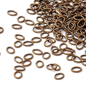 May include: A collection of small, oval-shaped, antique bronze-colored jump rings scattered on a white surface. The rings are open, ready for use in jewelry making or other crafts. The metal has a slightly textured appearance.