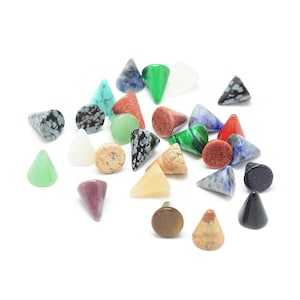 May include: A collection of gemstone plugs in various shapes and colors, including green, red, blue, and speckled patterns. The plugs are cone-shaped, round, and triangular, showcasing a variety of natural stone textures and hues. The plugs are scattered on a white surface.