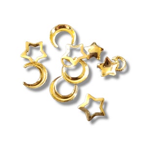 May include: Gold-colored celestial-themed charms. The charms include open star shapes and crescent moons. The charms are scattered on a white background. These charms are suitable for jewelry making or other craft projects.
