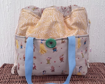 May include: A beige tote bag with a light blue handle and a green button closure. The exterior features a print of cartoon characters, while the interior lining has a yellow floral pattern. The bag is open, revealing the patterned interior.