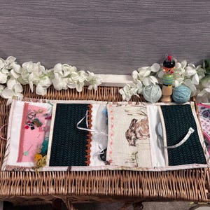 May include: A multi-compartment fabric organizer with a pink, green, and beige design. The organizer has a pocket for needles, a pocket for small tools, a pocket for thread, and a pocket for a tape measure. The organizer is sitting on a wicker basket.