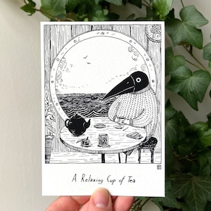 Könnte beinhalten: Schwarz-Weiß-Illustration einer krähenartigen Figur in einem Pullover, die Tee an einem Bullauge mit Meerblick genießt. Eine Teekanne, Snacks und der Text "A relaxing cup of tea" befinden sich auf dem Tisch. Das Kunstwerk ist auf einer weißen Karte.